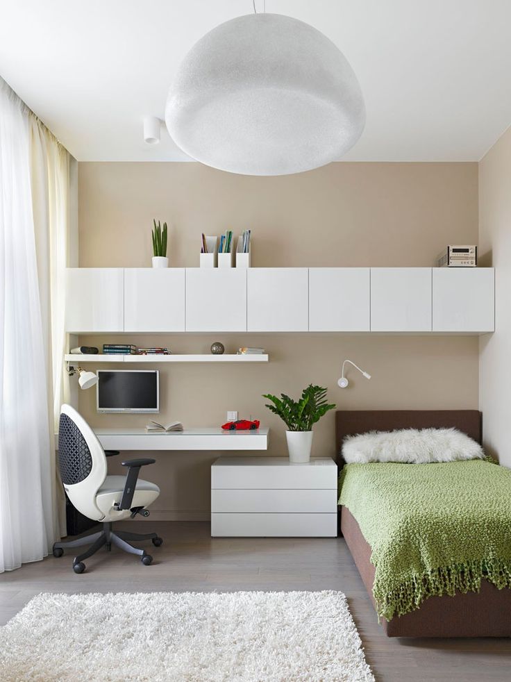 Small bedroom with tan walls, white floating cabinets and desk, brown bed with green throw, white round pendant light